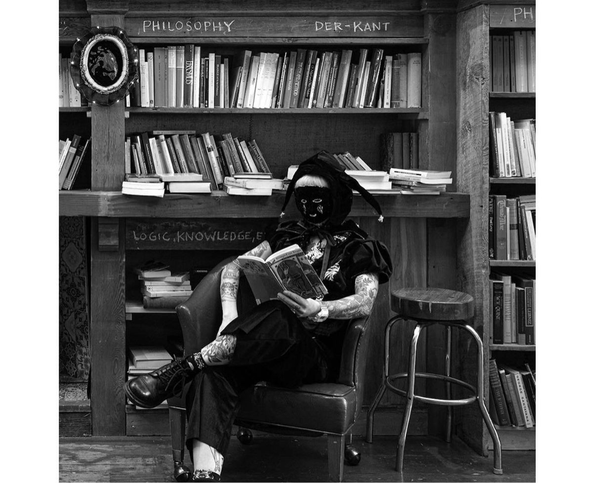 A beautiful young woman dressed like a traditional jester in a handmade outfit reading a book in the philosophy section of a bookstore